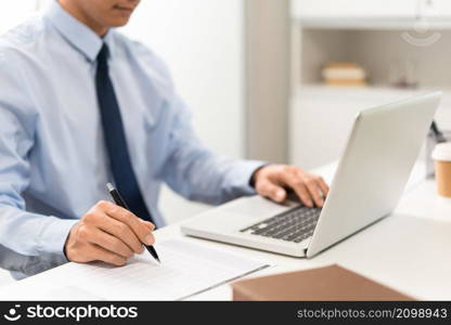 Working Man Conept The male officer sitting in the office, laying his left hand on the laptop, and writing something on the paper with his right hand.