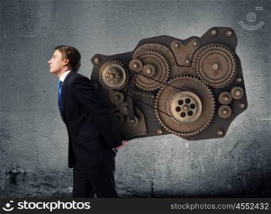 Working mechanism. Young businessman carrying cogwheel mechanism on back