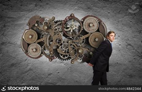 Working mechanism. Young businessman carrying cogwheel mechanism on back