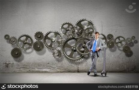 Working mechanism. Young determined businessman leaning on wrench and cogwheels at background