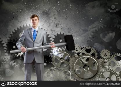 Working mechanism. Young determined businessman with hammer and cogwheels at background