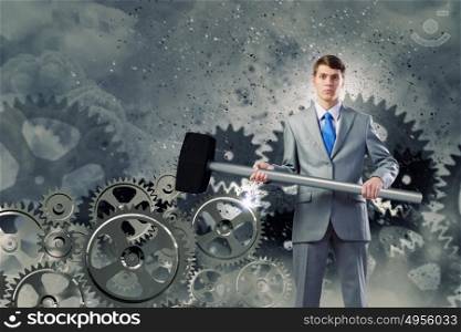 Working mechanism. Young determined businessman with hammer and cogwheels at background