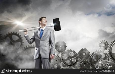 Working mechanism. Young determined businessman with hammer on shoulder and cogwheels at background