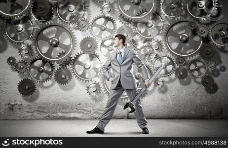 Working mechanism. Young determined businessman with wrench in hands and cogwheels at background