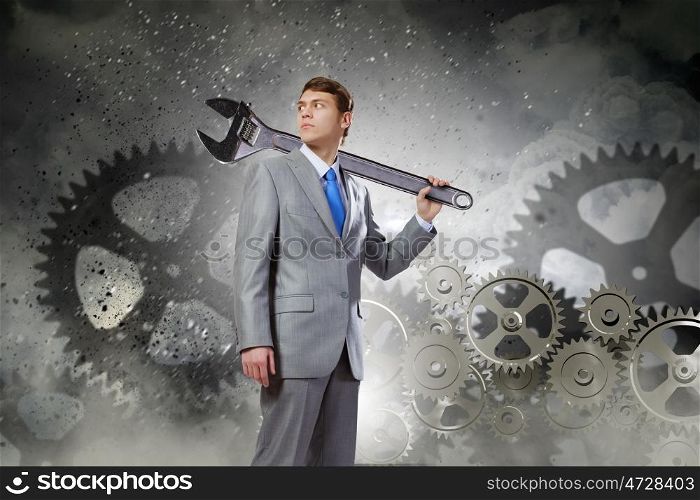 Working mechanism. Young determined businessman with wrench on shoulder and cogwheels at background