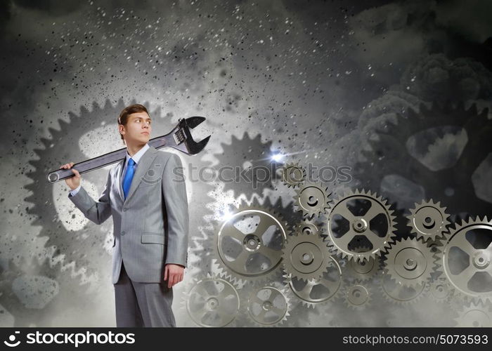 Working mechanism. Young determined businessman with wrench on shoulder and cogwheels at background