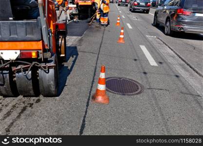 Working team patches the damaged part of the asphalt on the fenced cones stretch of city street, image with copy space.. Road orange cones enclose the work crew and road equipment from the carriageway