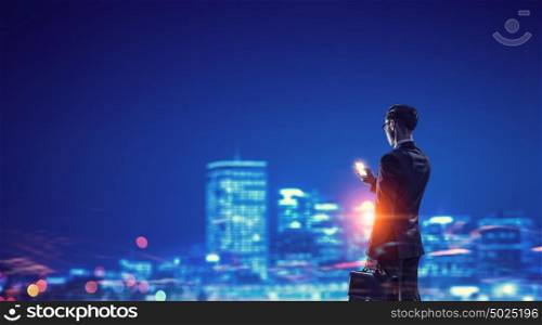 Working till late night. Young businessman with mobile phone against night city background