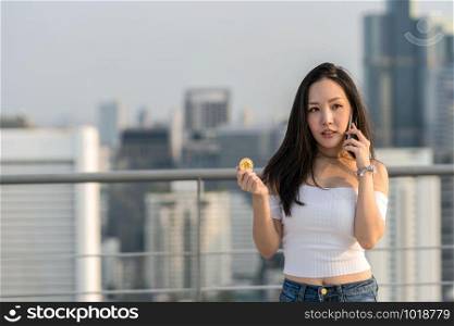Working woman in casual suit holding bitcoin and using the smart mobile phone over the cityscape blurred background, business and cryptocurrency concept