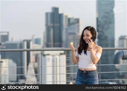 Working woman in casual suit holding bitcoin and using the smart mobile phone over the cityscape blurred background, business and cryptocurrency concept