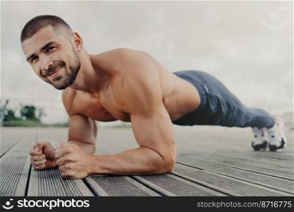 Workout and healthy lifestyle concept. Pleased muscular bearded young man stands in plank pose, makes sport training outdoors, looks with determined expression, concentrated on getting better