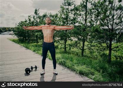Workout, healthy lifestyle and bodybuilding concept. Strong muscular European man stretches resistance band, wears sneakers, stands outdoor, enjoys exercising in open air, has athletic body.