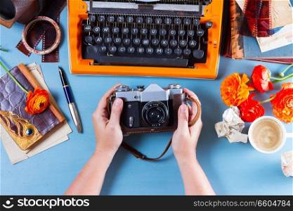 Workspace with someone hands holding old photo camera, on table with orange vintage typewriter, flat lay scene. Workspace with vintage orange typewriter