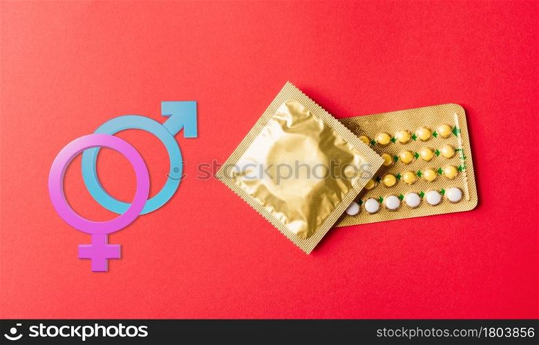 World sexual health or Aids day. Male, female gender signs condom on wrapper pack and contraceptive pills blister hormonal birth control pills, studio shot isolated on a red background