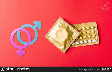 World sexual health or Aids day. Male, female gender signs condom on wrapper pack and contraceptive pills blister hormonal birth control pills, studio shot isolated on a red background