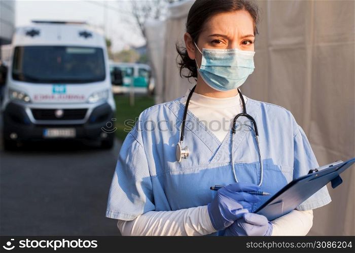 Worried & anxious UK NHS EMS doctor,holding patient report form folder,standing in front of ambulance car,tired & exhausted from working long shifts,outdoor PPE portrait,Coronavirus COVID-19 pandemic