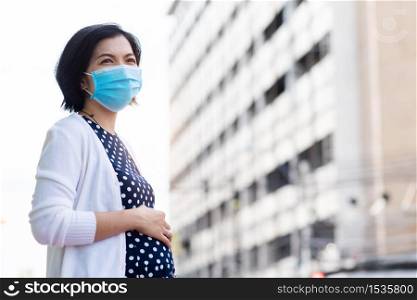 Worried Asian pregnant woman in protective face mask for air pollution and pandemic coronavirus or covid-19 touching her belly and standing against building background at outdoor with new normal life