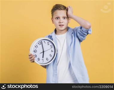 worried boy with hand his head holding white clock standing against yellow background