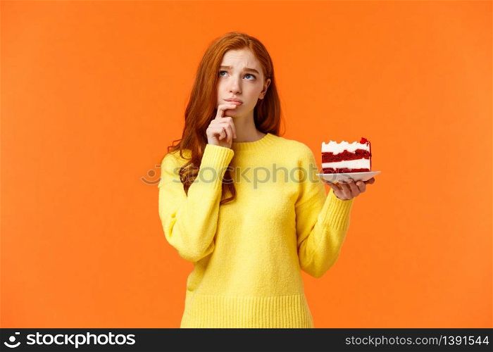 Worried, concerned silly young redhead girl taking care her body, calculating calories, being diet and want eat delicious cake, holding piece of dessert, look up thoughtful and troubled, orange wall.. Worried, concerned silly young redhead girl taking care her body, calculating calories, being diet and want eat delicious cake, holding piece of dessert, look up thoughtful and troubled, orange wall