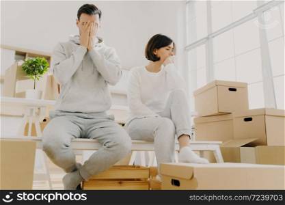 Worried couple pose near stack of cardboard boxes, have to move in other flat, have no money for rent, have dejected sad expressions, sit together in white room, have family conflict and divorce