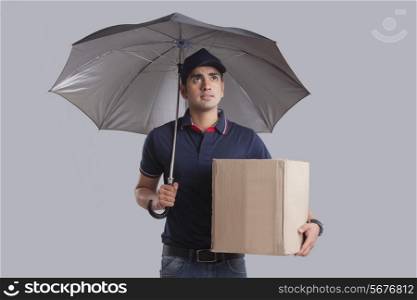 Worried delivery man with package and umbrella over gray background