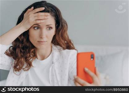 Worried girl is reading trouble news looking at smartphone screen. Young attractive woman is chatting on phone or browsing internet at home. European girl is getting stress and anxiety attack.. Worried girl is reading trouble news looking at smartphone screen. Stress and anxiety attack.