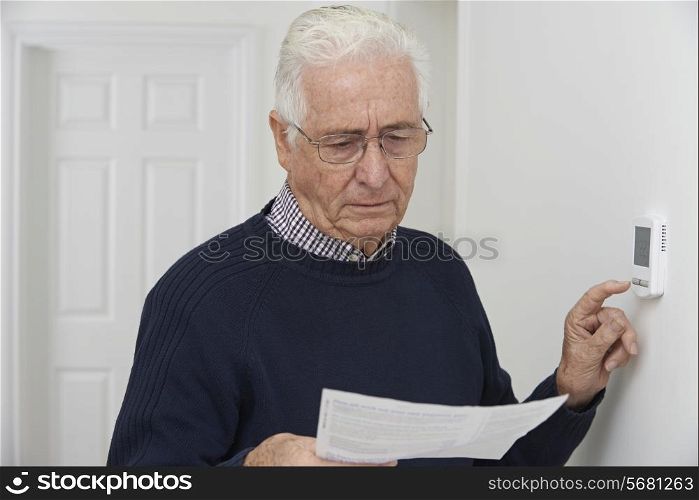 Worried Senior Man With Bill Turning Down Central Heating Thermostat