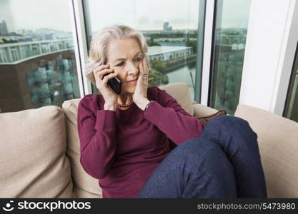 Worried senior woman answering smart phone on sofa at home