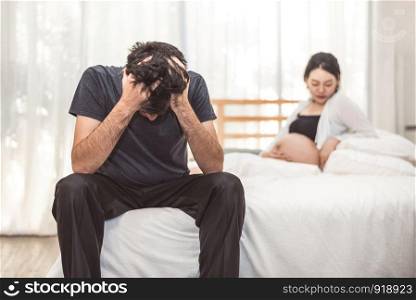 Worried stress man sitting on bed with hand on forehead in bedroom in serious mood emotion with pregnant wife woman background. Major Depressive Disorder called MDD concept. Physical healthcare