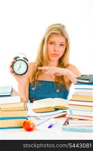 Worried teen girl sitting at table with books and pointing finger on alarm clock&#xA;