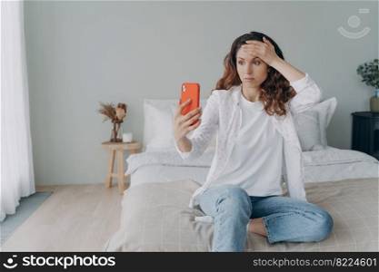 Worried tired woman feeling stress, holding smartphone having video call sitting in bedroom. Upset female blogger streaming online in social networks dissatisfied with the number of viewers.. Worried tired young woman having video call, holding smartphone, feeling stress, sitting in bedroom