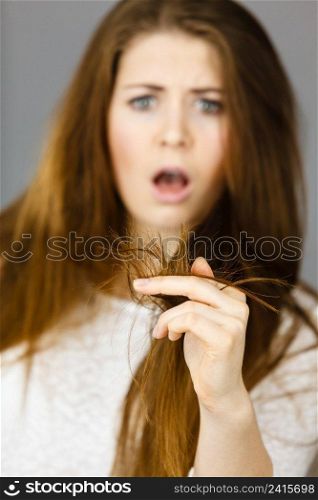 Worried woman looking at her dry split long brown hair ends, thinking of good treatment. Haircare and hairstyling concept.. Worried woman looking at her dry hair ends