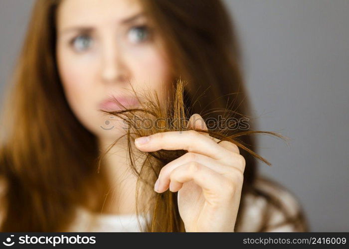 Worried woman looking at her dry split long brown hair ends, thinking of good treatment. Haircare and hairstyling concept.. Worried woman looking at her dry hair ends