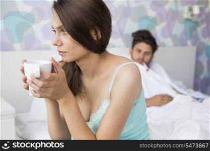 Worried young woman holding coffee mug while ill man relaxing in background