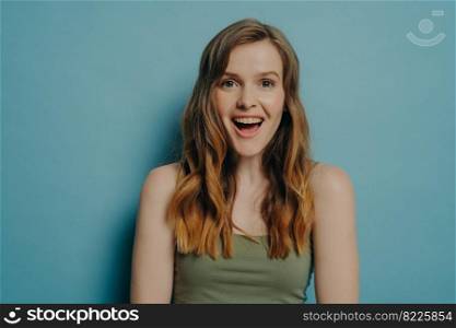 Wow emotion. Studio portrait of amazed astonished young female in tank top expressing happiness and excitement, looking at camera with surprised face expression, standing isolated on blue background. Amazed astonished young female in tank top expressing happiness and excitement, isolated on blue