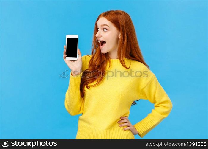 Wow look at this app. Amused and wondered, excited redhead girl looking at smartphone display, showing mobile screen, shopping site or application, standing blue background. Advertisement concept. Wow look at this app. Amused and wondered, excited redhead girl looking at smartphone display, showing mobile screen, shopping site or application, standing blue background