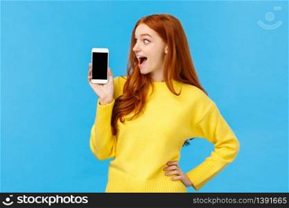 Wow look at this app. Amused and wondered, excited redhead girl looking at smartphone display, showing mobile screen, shopping site or application, standing blue background. Advertisement concept. Wow look at this app. Amused and wondered, excited redhead girl looking at smartphone display, showing mobile screen, shopping site or application, standing blue background