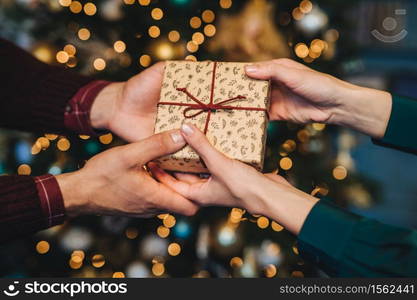 Wrapped box in woman`s and man`s hands. Affectionate husband congratulates his wife with New Year, presents her holiday gift box, stand against decorated Christmas tree. Holiday concept