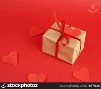 wrapped square box with a gift in brown craft paper and tied with a thin silk red ribbon on a red background, empty paper heart on a gift