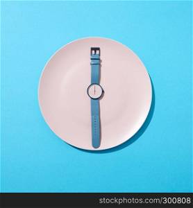 Wristwatch with six o'clock and blue bend on a white plate on a blue background. Time to lose weight, eating control or diet concept. Top view.. Watch with time six o'clock on a white plate on a blue background. Concept of limiting the intake of food diet and weight loss. Flat lay.