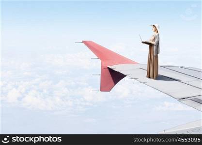 Writer woman working in isolation. Woman in dress and hat standing on airplane wing and working on laptop