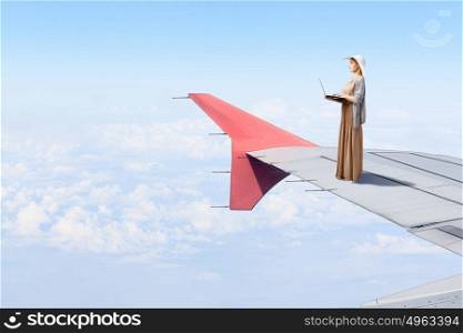 Writer woman working in isolation. Woman in dress and hat standing on airplane wing and working on laptop