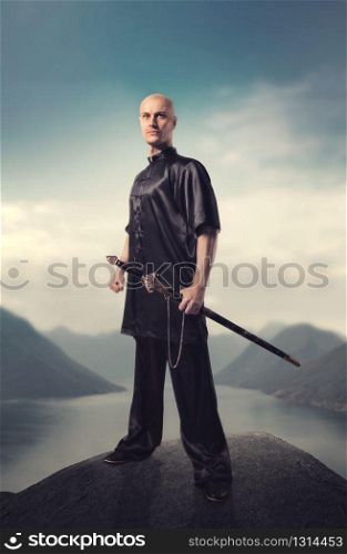 Wushu master with sword poses on the top of mountain, martial arts. Man in black cloth poses with blade