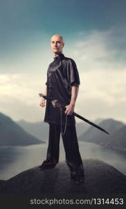 Wushu master with sword poses on the top of mountain, martial arts. Man in black cloth poses with blade