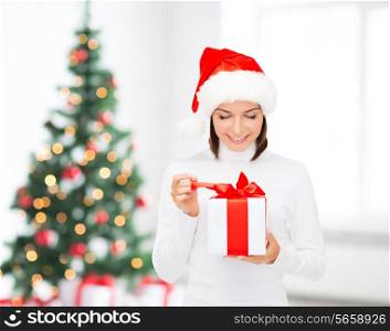 x-mas, winter, happiness, holidays and people concept - smiling woman in santa helper hat with gift box over living room and christmas tree background