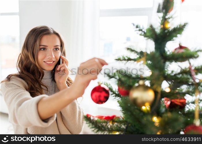 x-mas, winter holidays and people concept - happy young woman decorating christmas tree and calling on smartphone at home