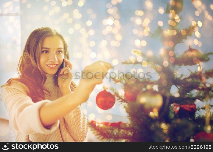 x-mas, winter holidays and people concept - happy young woman decorating christmas tree and calling on smartphone at home. woman with smartphone decorating christmas tree