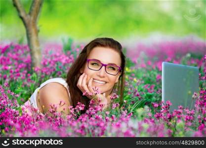 &#xA;Portrait of beautiful smiling student girl lying down on flowers meadow in the university garden, with pleasure working on laptop outdoors, using wireless internet
