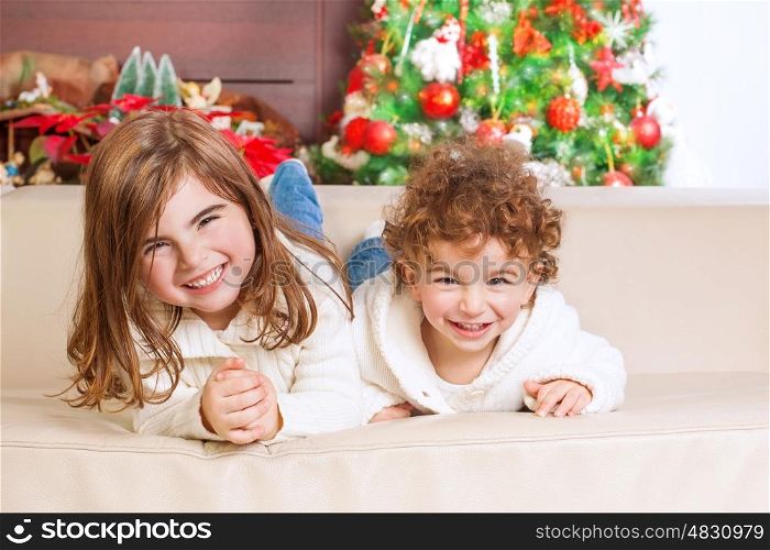 &#xA;Two happy kids lying down on sofa under beautiful decorated Christmas tree and laughing, cheerful family spending winter holidays at home