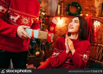 Xmas celebration, male person makes gift to beautiful young woman, fir-tree with decoration on background. Happy love couple celebrate christmas holidays